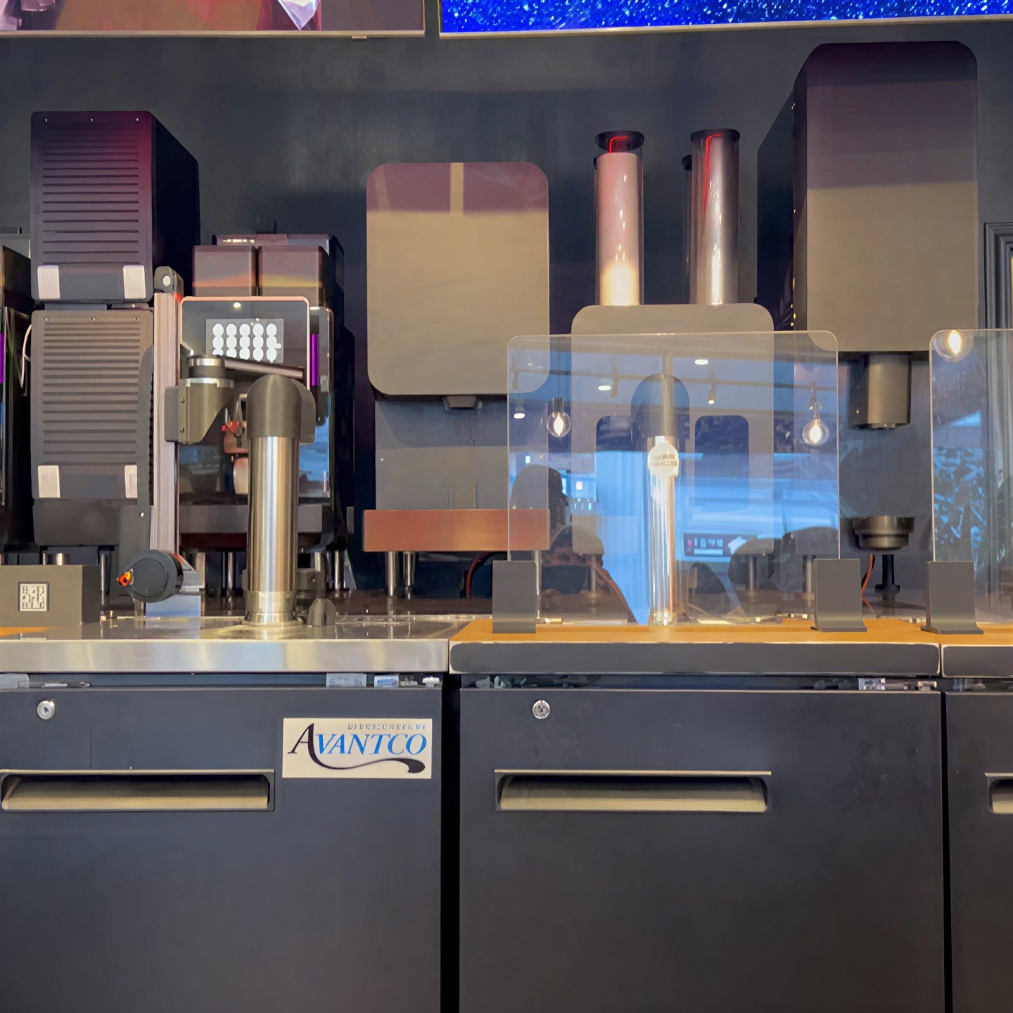 Machine bay close-up: stainless dispensing column, acrylic guard, undercounter refrigeration
