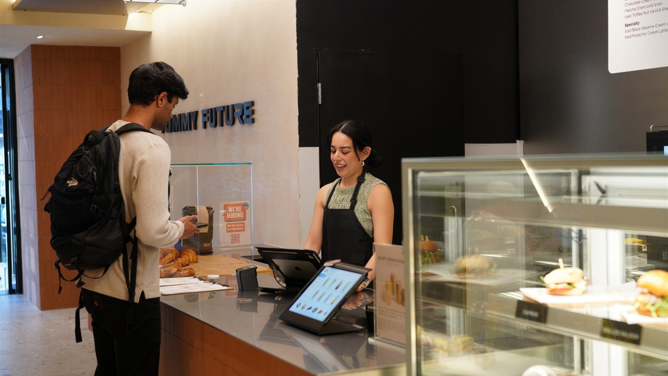 A customer with a backpack orders at the counter while a smiling staff member takes the order on an iPad point-of-sale; a WE'RE HIRING card stands on the counter wall behind her