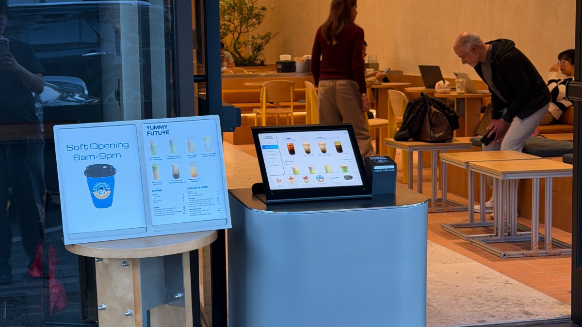 Store doorway with a Soft Opening sign, a menu board and an order screen, customers seated inside